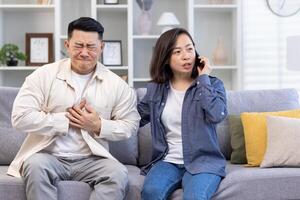 Young Asian family sitting on sofa at home. A man holds his hand to his chest, heart attack, stroke, panic attack, feels severe pain. A shocked woman calls an ambulance and calls a doctor. emergency call. photo