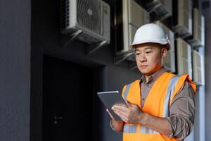 Serious Asian man works in construction and manufacturing. Standing outside in a hard hat and vest, holding a tablet, checking data, doing logistics and transportation. photo