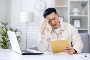 Upset and disappointed man in home office received message mail envelope with bad news, asian man working in home office at desk with laptop. photo