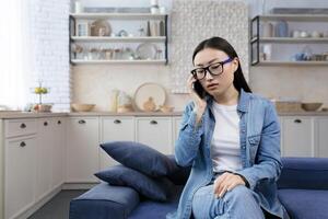 trastornado joven hermosa asiático mujer en lentes hablando en el teléfono sentado en el sofá foto