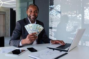 Portrait of an African American male businessman working in the office at a laptop and holding cash money in his hands, pointing and smiling at the camera. photo