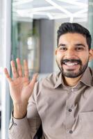 confidente joven indio masculino empresario hablando en un teléfono inteligente y participación arriba su mano como un saludo mientras en pie en un bien iluminado oficina ambiente. foto