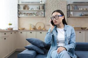 Worried and confused young Asian woman sitting on sofa at home and calling emergency service on mobile. Talks on the phone, asks for help. photo