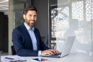 Portrait of a young successful businessman, executive director in a suit working in the office at the table on a laptop, looking and smiling at the camera. photo