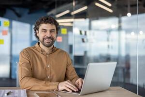 Confident male professional, identified as Hispanic, engaging with a computer in a modern office setting while looking at the camera. photo