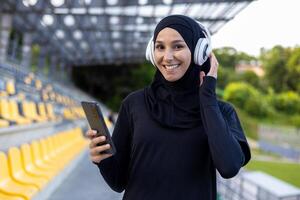 retrato de un joven hijab musulmán mujer en un Deportes estadio corriendo y haciendo activo físico ejercicios, sonriente y mirando a el cámara, utilizando el teléfono y auriculares a escucha a música. foto