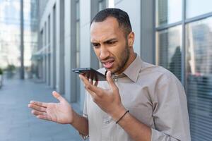 Angry businessman in shirt recording audio message from outside office building, african american man using assistant giving commands to artificial intelligence using smartphone. photo