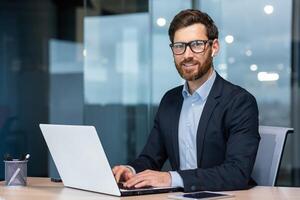 Portrait of mature successful and happy businessman, senior man with beard smiling and looking at camera, investor working with laptop inside office in business suit. photo