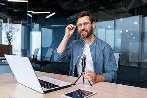 retrato de exitoso hombre dentro oficina, empresario con profesional micrófono grabación en línea formación curso y audio podcasts, utilizando ordenador portátil sentado a escritorio laboral, mirando a cámara. foto