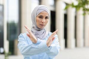 Empowered adult Muslim woman wearing a hijab crosses her arms in an 'X' gesture, signaling stop or denial outside. photo