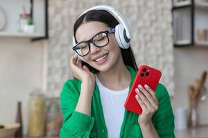 hermosa joven mujer en lentes y verde camisa relajante a hogar en cocina, escuchando música foto