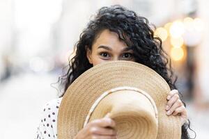 retrato de un hermosa juguetón latín americano mujer, un joven turista caminando mediante el ciudad en el noche, sonriente y mirando a el cámara, participación un Paja sombrero en su mano. foto