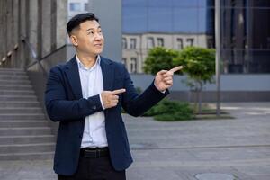 Asian businessman in suit pointing with both hands to side on a sunny day, standing outside with buildings in background. photo