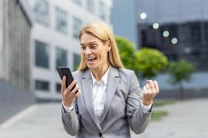 Successful mature joyful businesswoman received online notification message with good results achievement on phone, woman boss uses app on smartphone, walks outside office building in business suit. photo