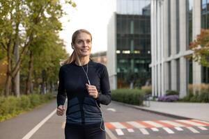 Mature experienced adult woman running in headphones in tracksuit outside modern building near trees, happy with workout sportswoman smiling active lifestyle. photo