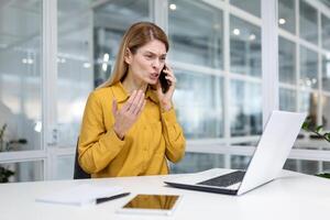 enojado y trastornado joven negocio mujer sentado en el oficina a el escritorio y hablando en el teléfono, resolviendo problemas y preguntas. foto