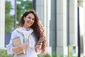 Joyful young woman with curly hair holding books and smartphone outside modern building, embodying success and happiness. photo