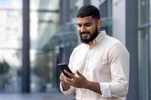 de cerca foto de un sonriente joven musulmán hombre en pie en un camisa cerca un oficina centrar y utilizando un móvil teléfono, hablando en un llamada