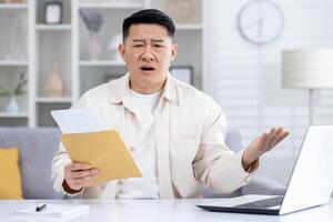 Worried korean male opening paper envelop with documents and sitting by desktop with disappointed facial expression. Mature businessman raising hands to camera as reaction for mail with bad news. photo