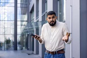 retrato de un joven árabe hombre en pie en el calle cerca un oficina edificio, participación un crédito tarjeta y un móvil teléfono, mirando preocupadamente a el cámara y extensión su manos foto