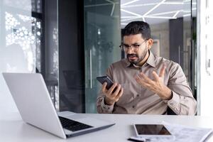 trastornado triste hombre a lugar de trabajo dentro oficina, empresario usos teléfono lee malo Noticias recibido por correo electrónico, empleado decepcionado sostiene teléfono inteligente en manos navega Internet sitios y social redes foto