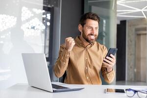 Man winner holding phone in hands, businessman at workplace happy with online achievement results, worker using app on smartphone, sitting inside office with laptop, wearing casual shirt. photo
