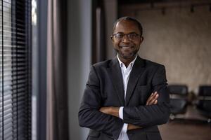 un profesional africano americano hombre en un traje soportes con confianza en un oficina configuración, brazos cruzado con un agradable sonrisa. foto