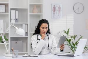 enfocado hembra médico en un blanco Saco sentado a su oficina escritorio, examinando médico información en un tableta con un ordenador portátil cerca en un bien iluminado clínica. foto