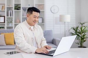 sonriente asiático hombre, empresario, persona de libre dedicación, estudiante trabajando estudiando a hogar utilizando computadora portátil, sentado a mesa y hablando en llamar, mecanografía. foto