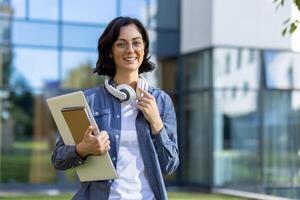 Portrait of young beautiful female student outside university campus, woman smiling and looking at camera wearing glasses, holding laptop on study pads. photo