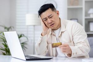 asiático hombre engañado por estafadores, hombre sentado a hogar a mesa participación banco crédito tarjeta, rechazado dinero transferir, negativo cuenta balance, trastornado y decepcionado. foto