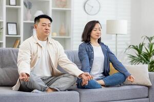 contento asiático familia a hogar en sofá meditando, Pareja hombre y mujer sentado en loto posición en vivo habitación con cerrado ojos descansando relajante juntos. foto