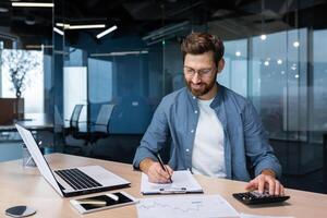 exitoso financiero inversor trabajos con documentos y cuentas dentro oficina, empresario con barba sonriente satisfecho con trabajo resultados y logros, hombre dentro oficina con ordenador portátil laboral. foto