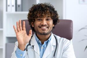 Webcam view, online call consultation, young Indian doctor in white medical coat looking at camera, waving hand greeting to patient, doctor working remotely inside clinic office with laptop. photo