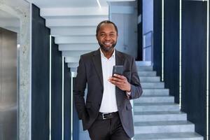 retrato de contento exitoso africano americano jefe, hombre sonriente y mirando a cámara, empresario participación teléfono utilizando en línea solicitud, hombre caminando abajo corredor de edificio dentro oficina. foto