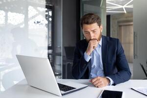 A young male businessman sits in the office at the desk, is sick and coughs, covers his mouth with his hand. photo