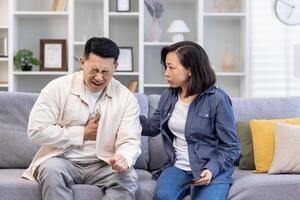 Young Asian family sitting on sofa at home. A man holds his hand to his chest, heart attack, stroke, panic attack, feels severe pain. Shocked woman tries to help, give first aid. photo