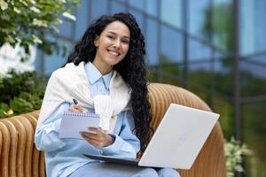 Successful diverse entrepreneur sitting on wooden bench and making notes in daybook while holding computer on laps. Smiling female writing to do list by hand for not skipping important tasks. photo