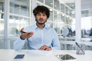 Young man with curly hair, speaking and gesturing, sits at a white desk in a modern office environment, giving a presentation. photo