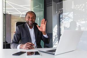 retrato de exitoso maduro africano americano jefe, hombre sonriente y mirando a cámara sentado a escritorio dentro oficina, empresario ondulación mano gesto de saludo y amistad. foto