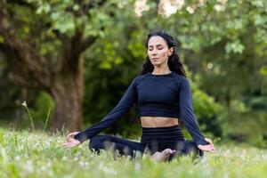 joven hermosa Hispano mujer en parque sentado en aptitud estera y meditando en loto pose, mujer descansando después aptitud y corriendo con ojos cerrado, respiración Fresco aire en chandal. foto