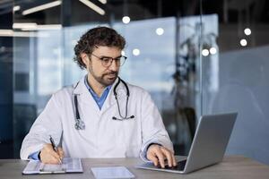 un profesional masculino médico en un laboratorio Saco utilizando un ordenador portátil y escritura notas en un portapapeles en un moderno cuidado de la salud ajuste. foto