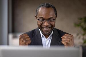 un lleno de alegría africano americano empresario celebra un exitoso acuerdo o logro mientras trabajando en su ordenador portátil. foto