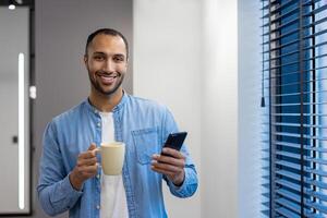 retrato de un sonriente musulmán hombre, un oficina trabajador y un persona de libre dedicación, en pie en el salón por el ventana, participación un taza de café y un móvil teléfono, mirando a el cámara. foto