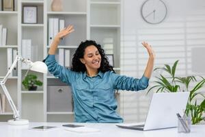 alegre mujer de negocios con brazos elevado en emoción a su escritorio en un moderno oficina, celebrando profesional logro. foto