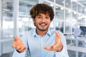 call online conference meeting, young successful hispanic man talking to colleagues remotely sitting in modern office, man looking at camera and smiling talking to colleagues in blue shirt. photo