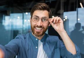 Portrait of successful businessman in casual shirt and glasses man looking at smartphone camera and smiling, camera view call talking with friends inside office remotely. photo