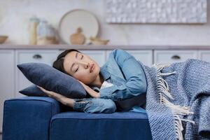 Close-up photo of a young beautiful Asian woman sleeping at home lying on the sofa covered with a blanket, resting after work for in the living room woman with closed eyes