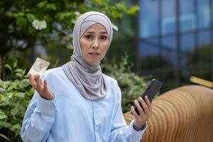 trastornado mujer en hijab mirando a cámara fuera de oficina edificio, negocio mujer participación banco crédito tarjeta y teléfono, rechazado dinero transferir, y en línea fraude foto