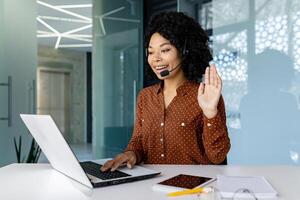 Woman with headset phone using laptop for remote communication and call, african american woman waving at camera greeting. photo